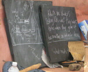two blackboards sitting outside (it appears) leaning against the side of a stucco house. One has drawings of a fireplace, perhaps planning on how to make it. The other has words, "Adults are always asking kids what they will do when they grow up, because they are looking for ideas"
